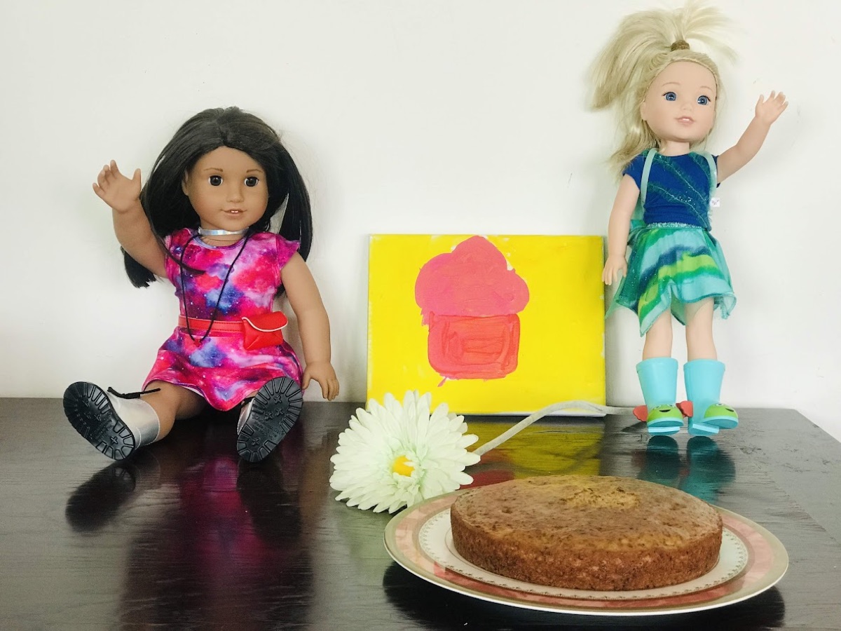 Two dolls, Lucy and Sophie, beside a homemade birthday cake and a white flower
