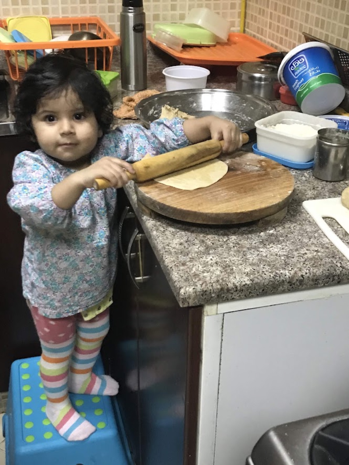 Vedhica as a toddler, standing on a stool and holding a rolling pin in the kitchen
